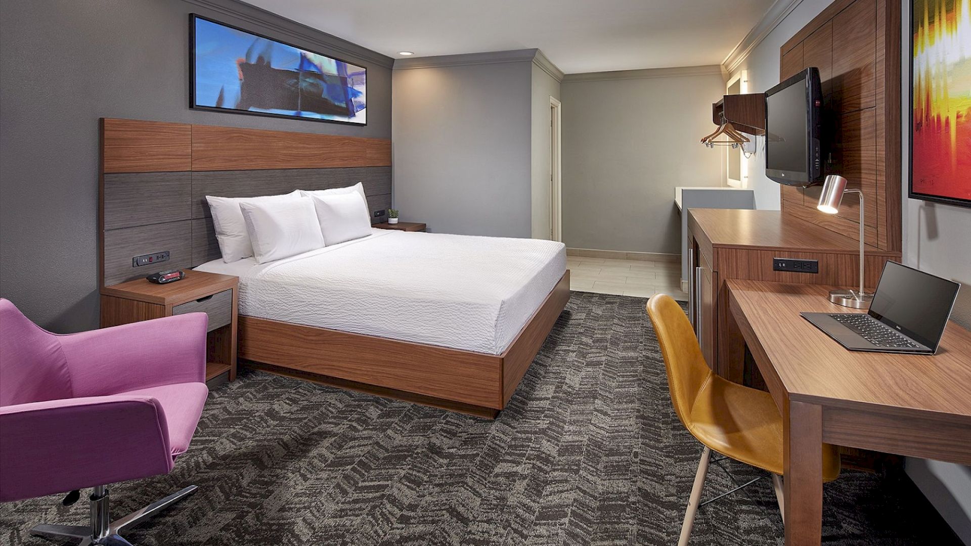 A modern hotel room features a double bed, purple chair, desk with yellow chair, wall-mounted TV, and colorful artwork on the walls.