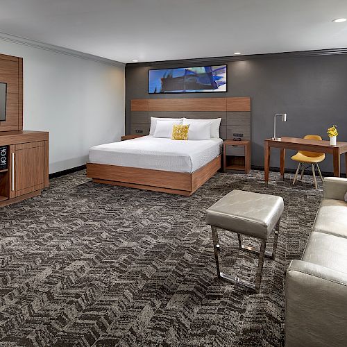 A modern hotel room with a large bed, TV, sofa, desk, and a patterned carpet, featuring contemporary decor and ample lighting.