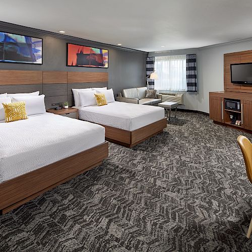 This image shows a hotel room with two beds, a TV, a desk with a chair, and a seating area near a window. The decor is modern and cozy.