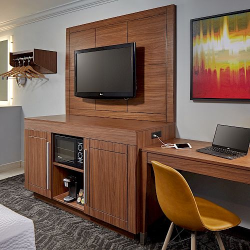 A hotel room with a TV on a wooden cabinet, a desk with a laptop and lamp, a yellow chair, and a framed colorful artwork on the wall.