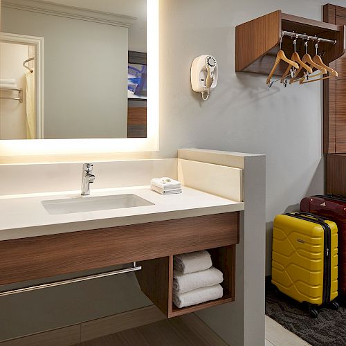 A hotel room with a vanity area, hanging clothes rack, TV, yellow and red suitcases, and towels below the sink, ending the sentence.