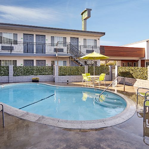 This image shows a hotel pool area with a few lounge chairs, a table with an umbrella, and buildings in the background ending the sentence.