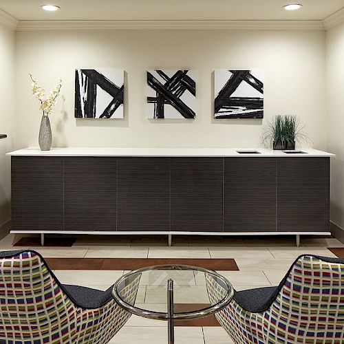 A modern waiting room with two patterned chairs, a glass table, abstract art, and decorative plants on a cabinet.