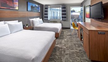 The image shows a modern hotel room with two beds, a large TV, a desk and chair, and a view of a balcony through sliding glass doors.