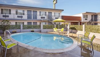 This image shows a motel courtyard with a small, kidney-shaped swimming pool, surrounded by green chairs and tables with yellow umbrellas.