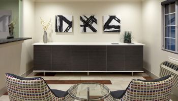 A modern living space with two patterned chairs, a small glass table, and a cabinet with abstract art and decorative items on top.