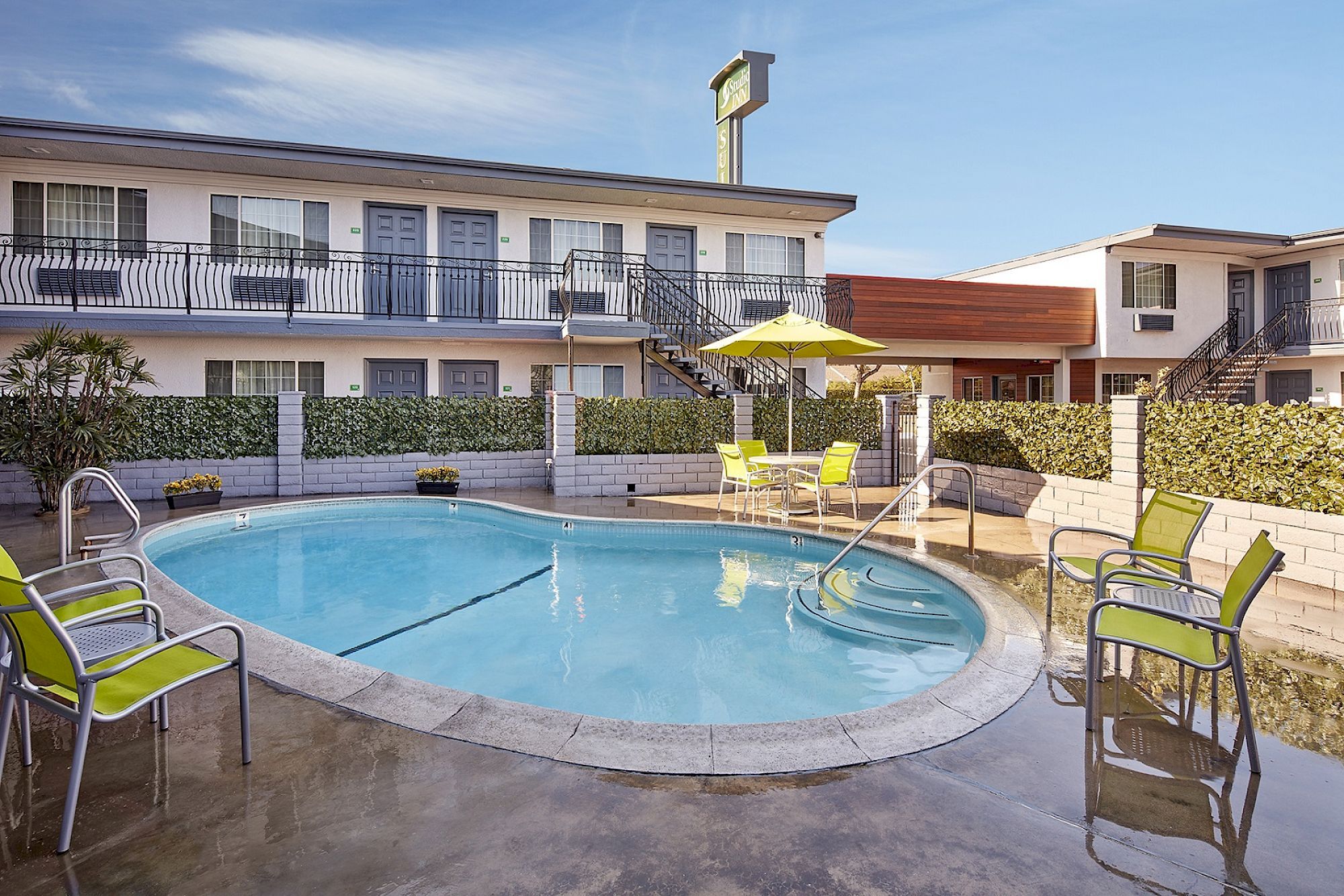 The image shows a motel courtyard with a kidney-shaped swimming pool, surrounded by green chairs and a table with a yellow umbrella.