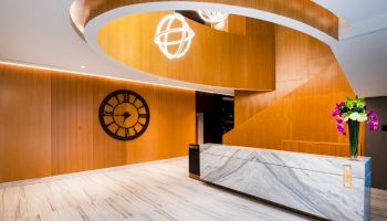 A modern reception area with a marble desk, wooden wall design, large wall clock, and decorative hanging lights, featuring a vase of flowers.