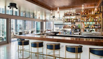 A modern bar with a wooden countertop, stocked shelves of alcohol, stylish bar stools, and large windows overlooking a cityscape is shown.