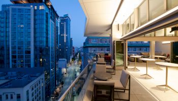 The image shows a modern rooftop terrace with seating, tables, and cityscape views at dusk. Urban buildings and street lights are visible.