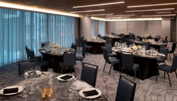 The image shows a banquet hall with round tables set for an event, decorated with white flowers and black napkins, under modern ceiling lights.