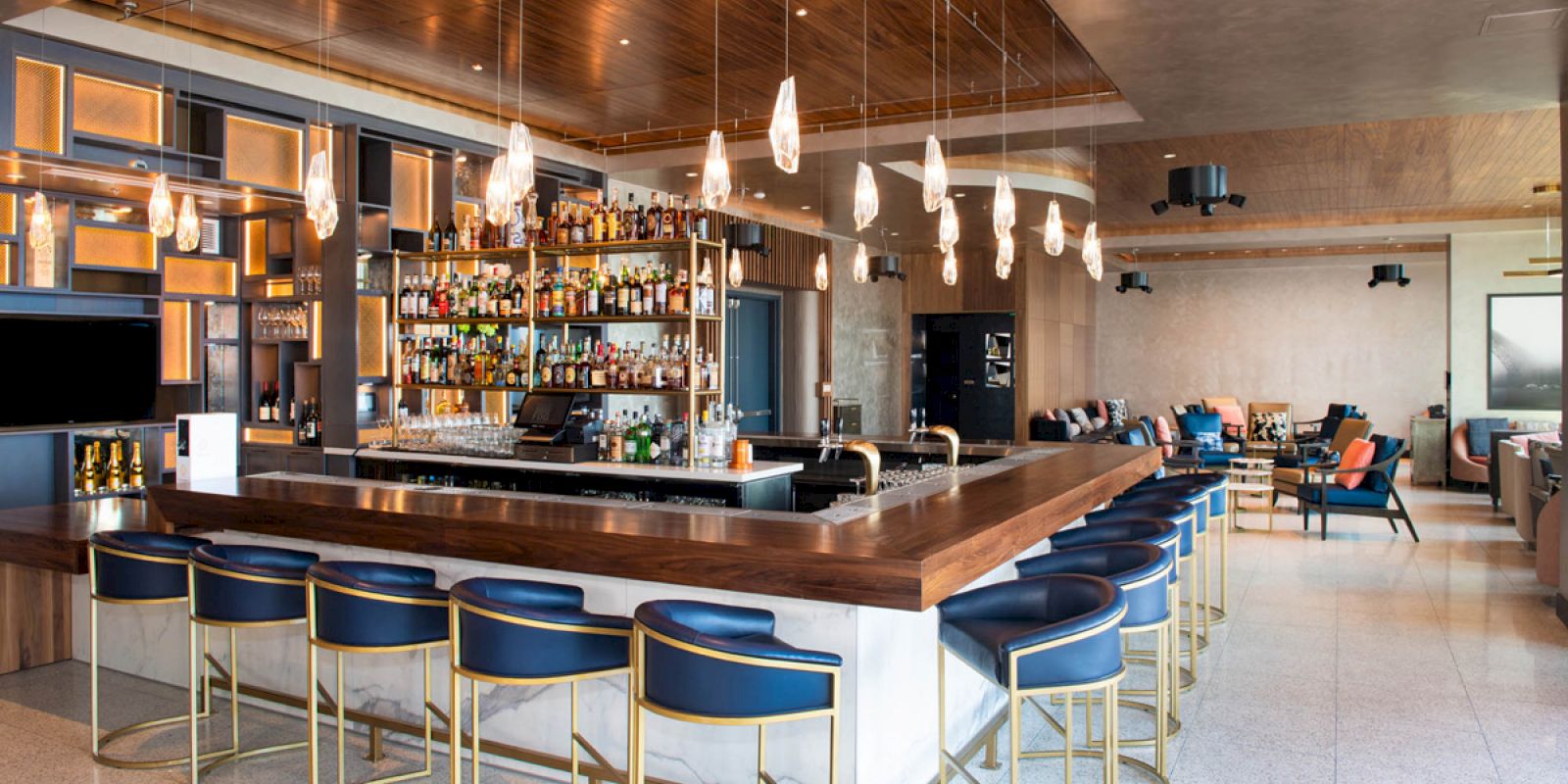 A modern bar with blue seating, wooden counter, hanging lights, and a fully stocked bar in a spacious, well-lit room with additional seating areas.