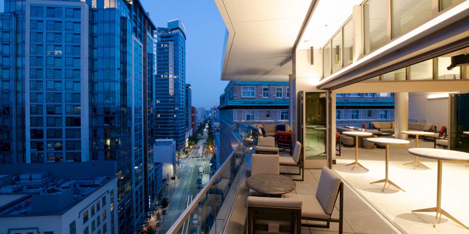 A modern urban balcony during twilight, with outdoor seating and tables, overlooking a city street lined with tall buildings.