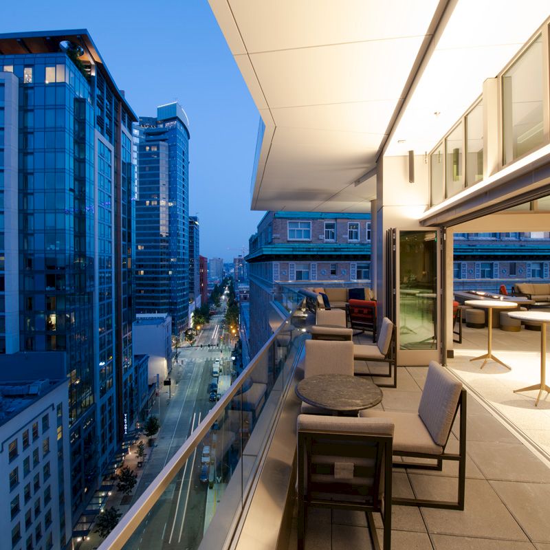 The image shows a modern balcony at twilight with seating and table arrangements, overlooking a cityscape of tall buildings and a street below.