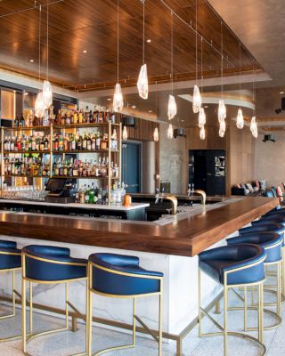 A modern bar with a wooden counter, blue bar stools, elegant hanging lights, and a variety of drinks displayed on shelves.