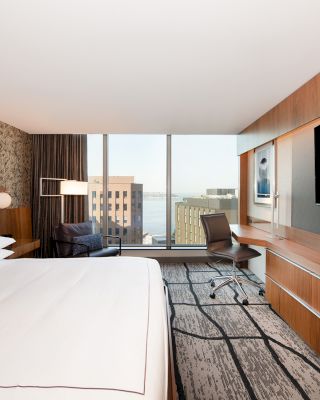 A modern hotel room with a king-size bed, large flat-screen TV, desk, chair, and a view of city buildings through a large window.