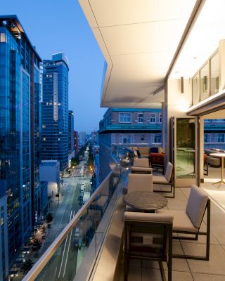 A modern cityscape at dusk with tall buildings and a well-furnished balcony featuring tables, chairs, and a view of the street below.