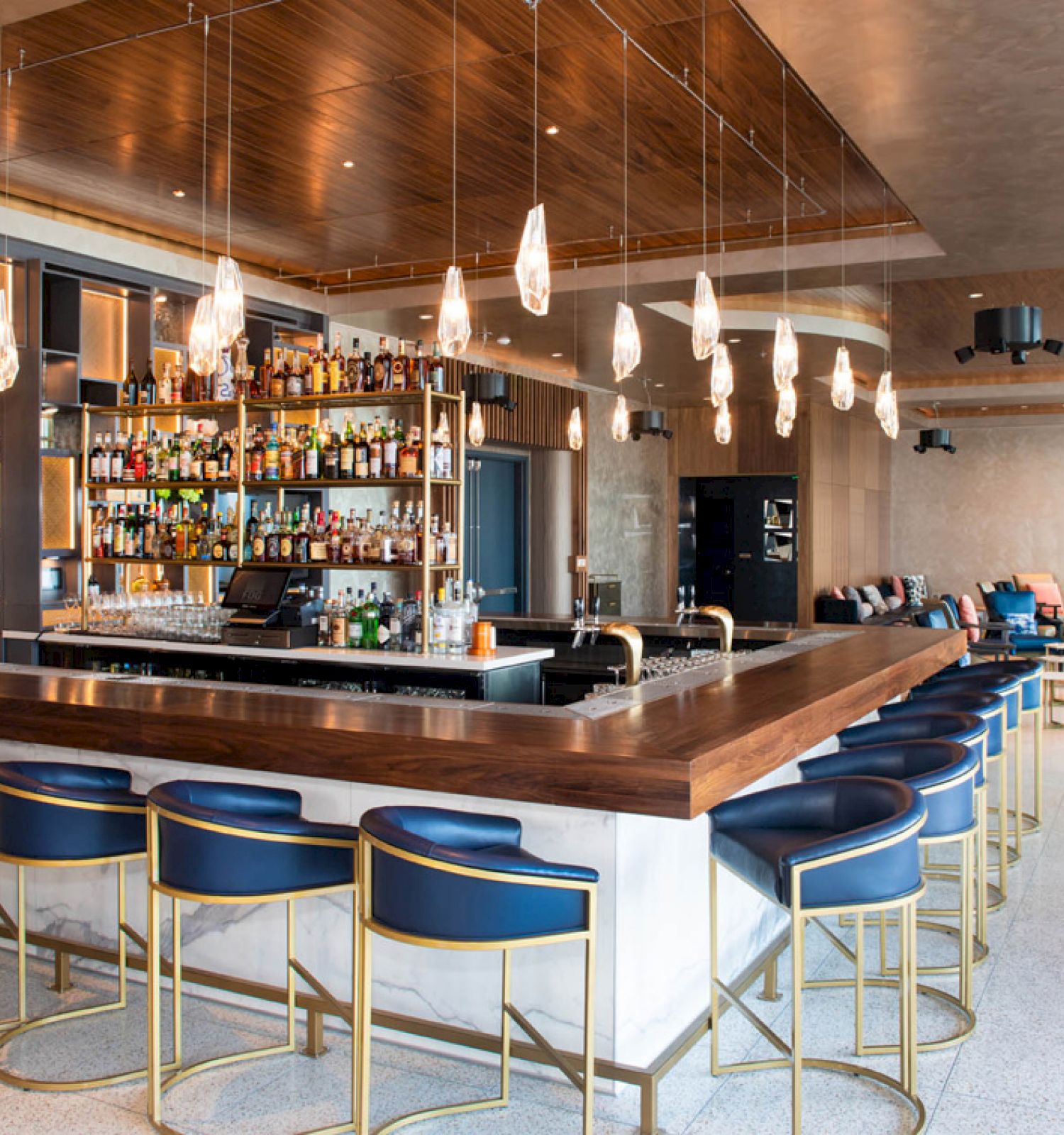 A modern bar with blue stools, wooden counters, a stocked liquor shelf, hanging lights, and a lounge area with seating.