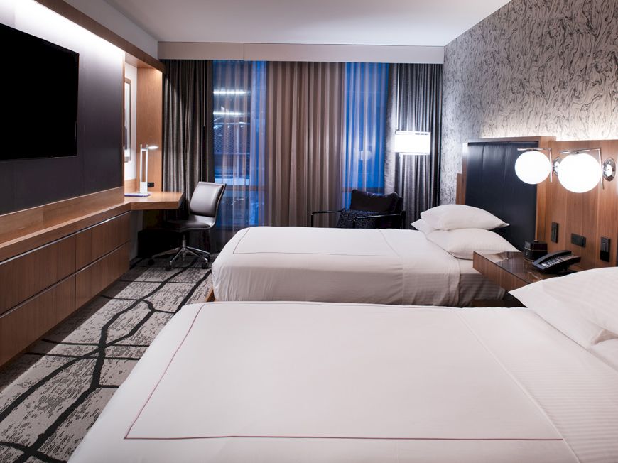 A modern hotel room with two beds, a flat-screen TV, a desk with a chair, and bedside lamps, featuring a patterned accent wall and floor-to-ceiling windows.
