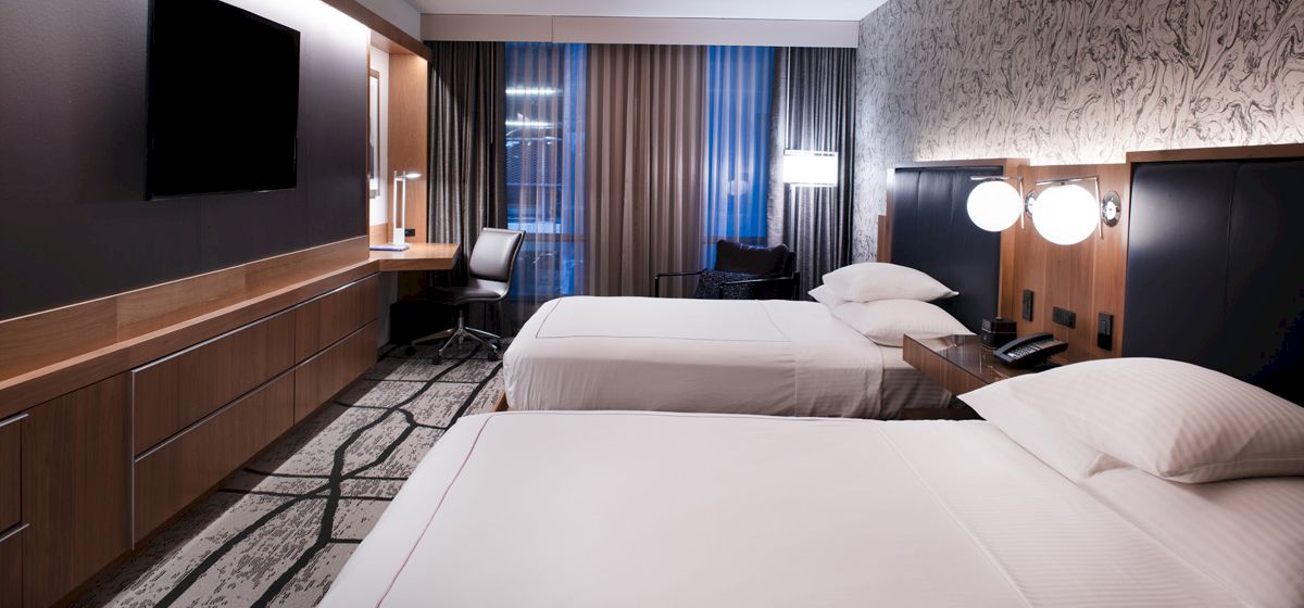 A modern hotel room with two beds, a flat-screen TV, a desk with a chair, and bedside lamps, featuring a patterned accent wall and floor-to-ceiling windows.