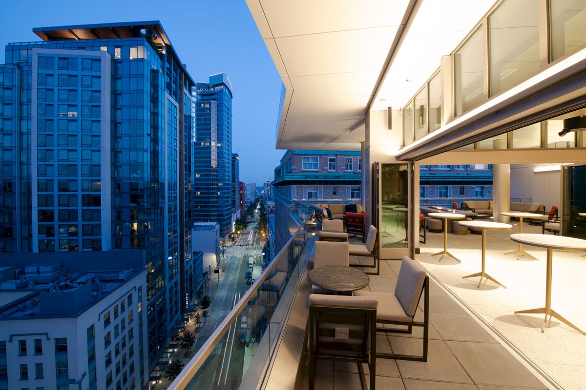A modern, outdoor balcony is attached to a building with tables and chairs, overlooking a city with tall buildings.
