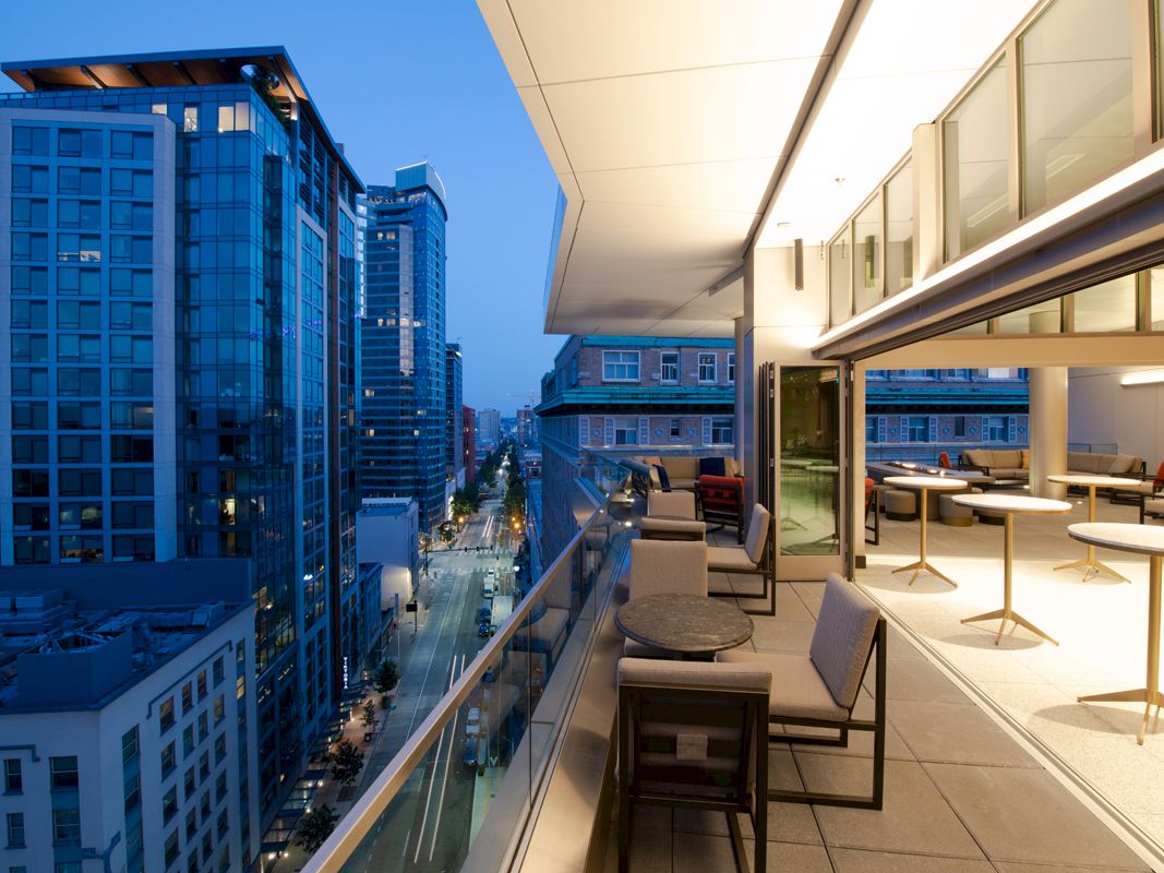 A modern, outdoor balcony is attached to a building with tables and chairs, overlooking a city with tall buildings.