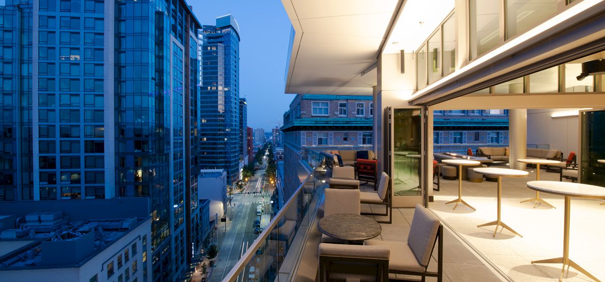 A modern, outdoor balcony is attached to a building with tables and chairs, overlooking a city with tall buildings.