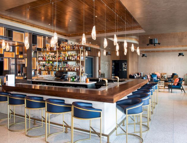 A modern bar with a wooden counter, blue barstools, hanging lights, and a well-stocked liquor shelf. There is seating in the background.