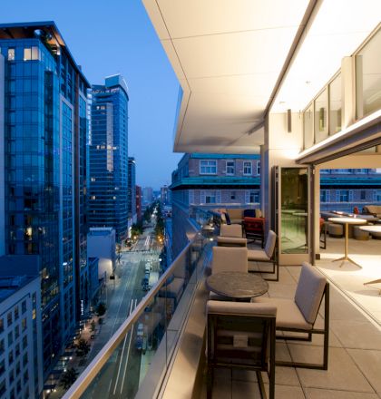 A modern outdoor balcony with seating and tables overlooking a cityscape of high-rise buildings at dusk.