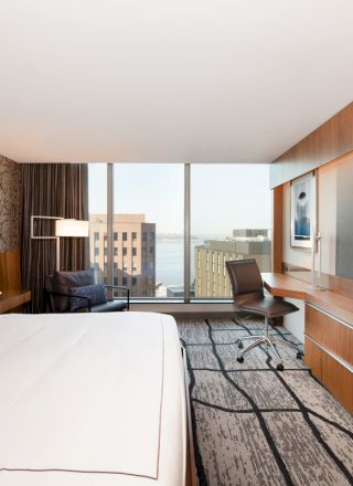 A modern hotel room with a large bed, a desk, a flat-screen TV, and a view of the city and water through floor-to-ceiling windows.