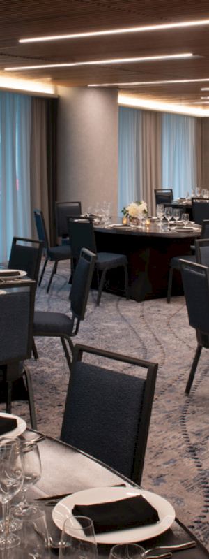 A banquet hall with round tables set for an event, featuring flowers, wine glasses, napkins, and black padded chairs under modern lighting.