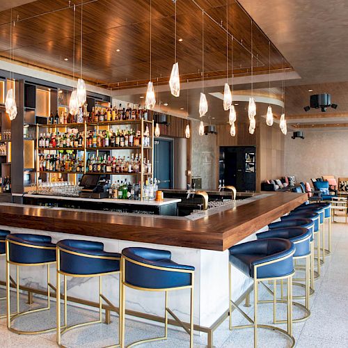 A modern bar with a wooden counter, blue bar stools, hanging lights, and a well-stocked liquor shelf. There's seating in the background.