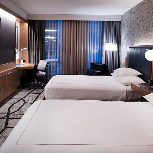 A modern hotel room with two double beds, a large TV, a desk with a chair, and stylish lighting and decor.