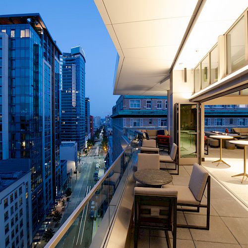 A modern high-rise balcony with patio furniture overlooks a city street lined with tall buildings at dusk, creating an urban twilight scene.