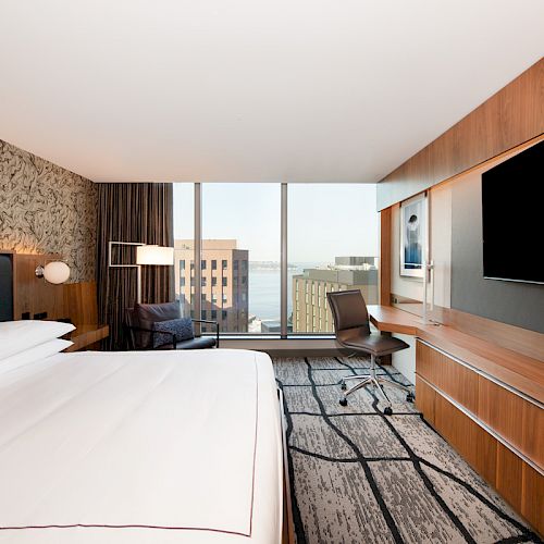 A modern hotel room with a large bed, wall-mounted TV, desk, and chair. It has a city view through a large window.