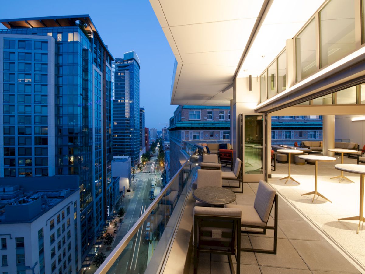 A modern balcony with tables and chairs overlooks an urban street lined with tall buildings at twilight.