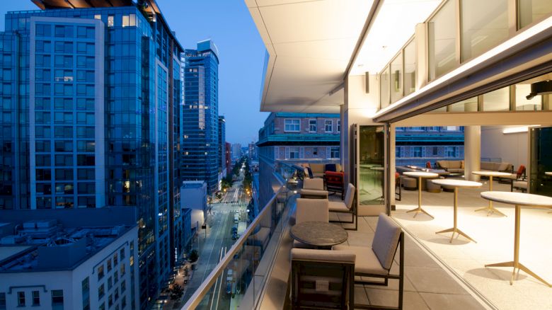 A modern balcony with tables and chairs overlooks an urban street lined with tall buildings at twilight.