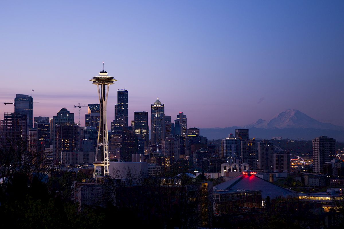 A twilight cityscape featuring a prominent needle-shaped tower and a skyline of tall buildings with a visible mountain range in the background.