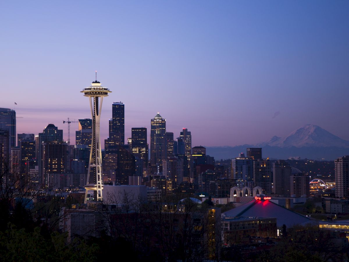 A twilight cityscape featuring a prominent needle-shaped tower and a skyline of tall buildings with a visible mountain range in the background.