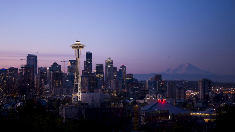 A twilight cityscape featuring a prominent needle-shaped tower and a skyline of tall buildings with a visible mountain range in the background.