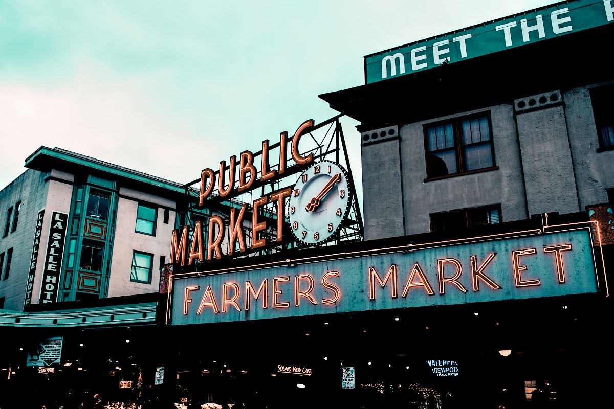 The image shows the iconic "Public Market" sign at a farmers market, with a clock and surrounding buildings. A neon "Farmers Market" sign is prominent.