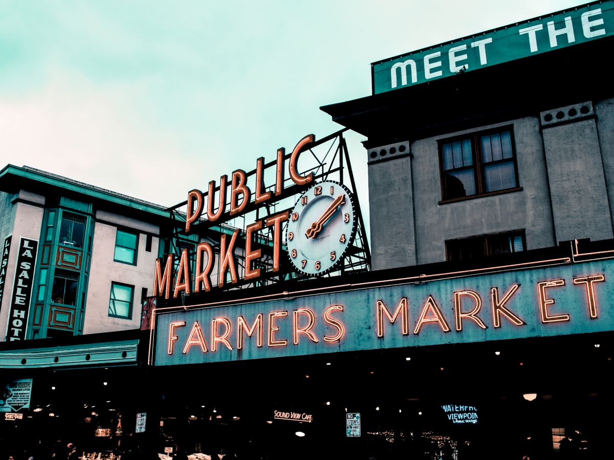 The image shows the iconic "Public Market" sign at a farmers market, with a clock and surrounding buildings. A neon "Farmers Market" sign is prominent.