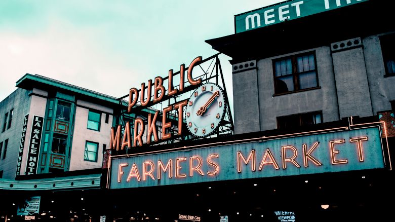 The image shows the iconic "Public Market" sign at a farmers market, with a clock and surrounding buildings. A neon "Farmers Market" sign is prominent.