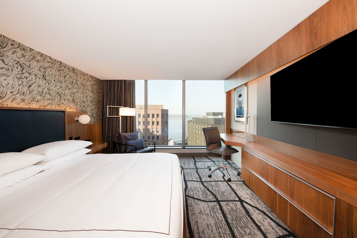 A modern hotel room features a large bed, a wall-mounted TV, a desk, a chair, and a window with a view of city buildings and water.