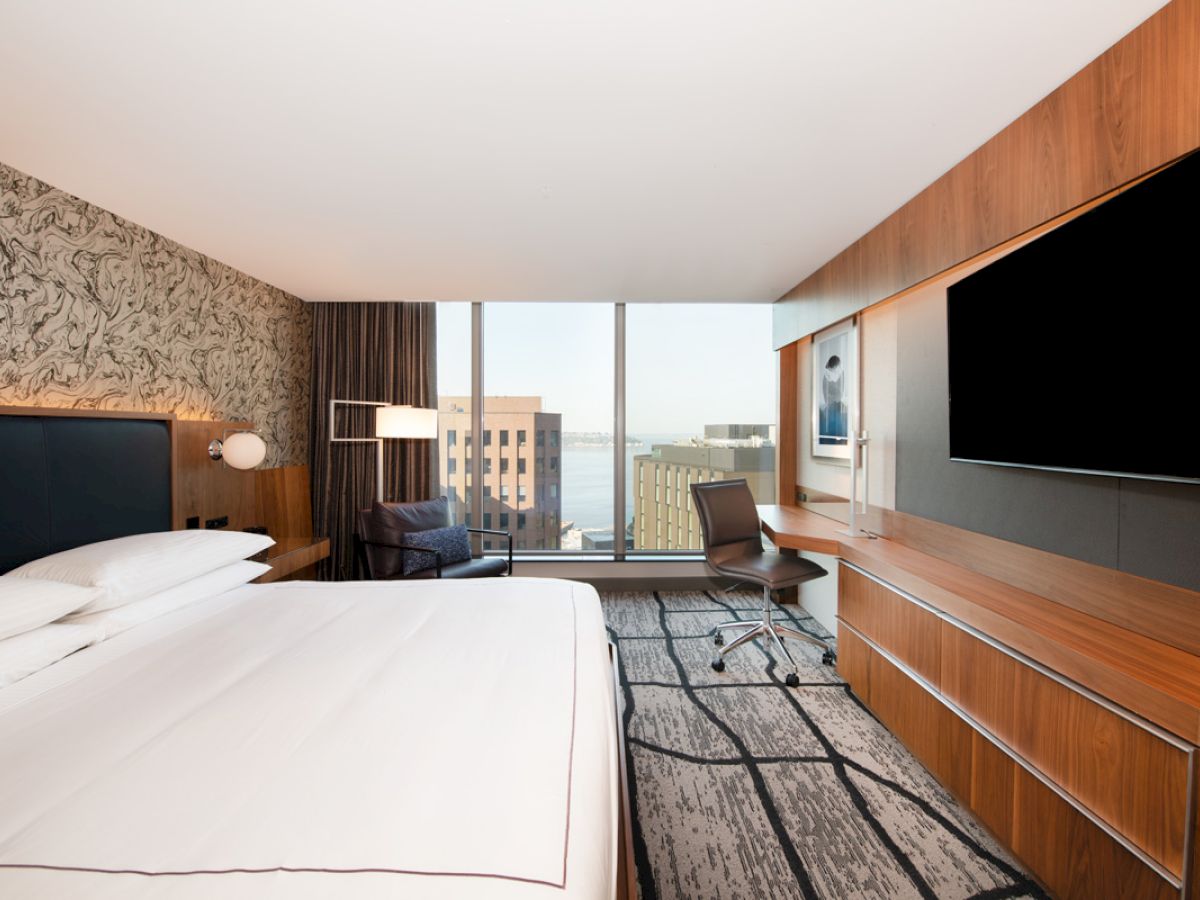 A modern hotel room features a large bed, a wall-mounted TV, a desk, a chair, and a window with a view of city buildings and water.