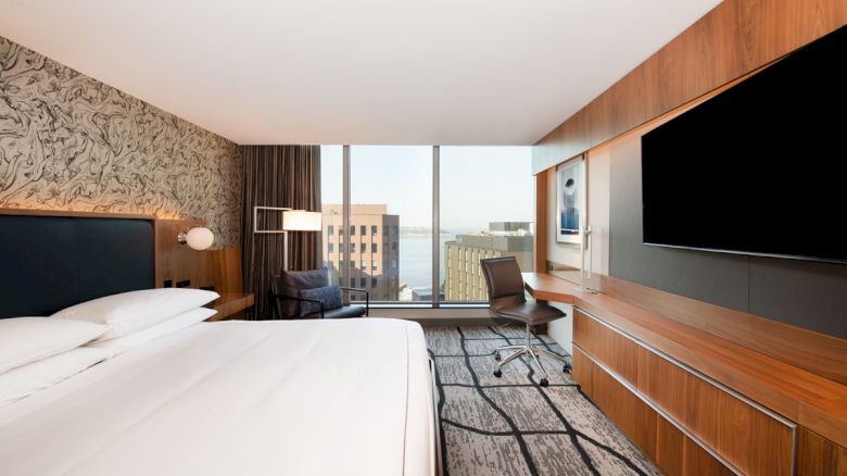 A modern hotel room features a large bed, a wall-mounted TV, a desk, a chair, and a window with a view of city buildings and water.