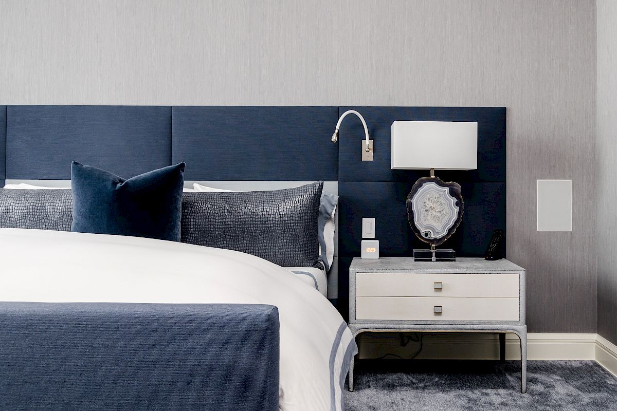 The image shows a modern bedroom with a blue upholstered bed, white bedding, a nightstand with a lamp, and decorative pieces on a grey carpeted floor.