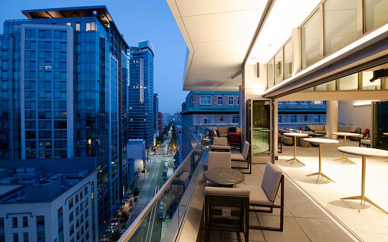 A modern balcony with seating and tables overlooking a city filled with tall buildings during the evening.