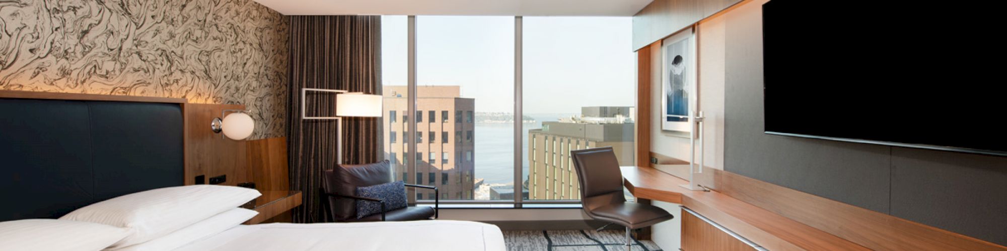 A modern hotel room with a large bed, wooden furnishings, a desk, a chair, and a large TV. The window offers a cityscape view.