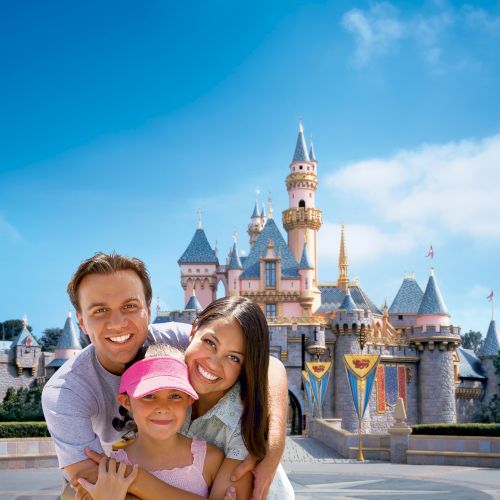 A family of three smiling, posing in front of a castle at a theme park. The child is wearing a pink cap. The sky is clear and blue.